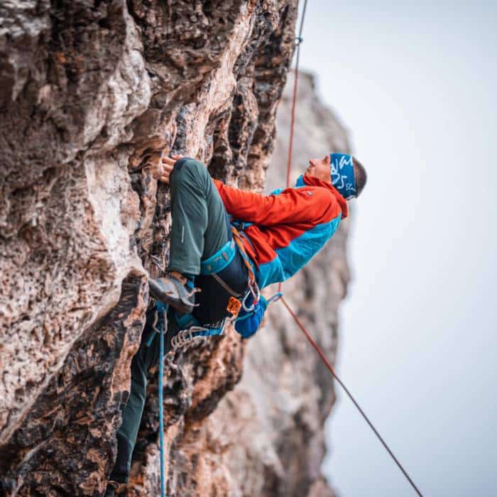 A climber in orange and blue gear scaling a steep, rocky cliff with a safety rope.